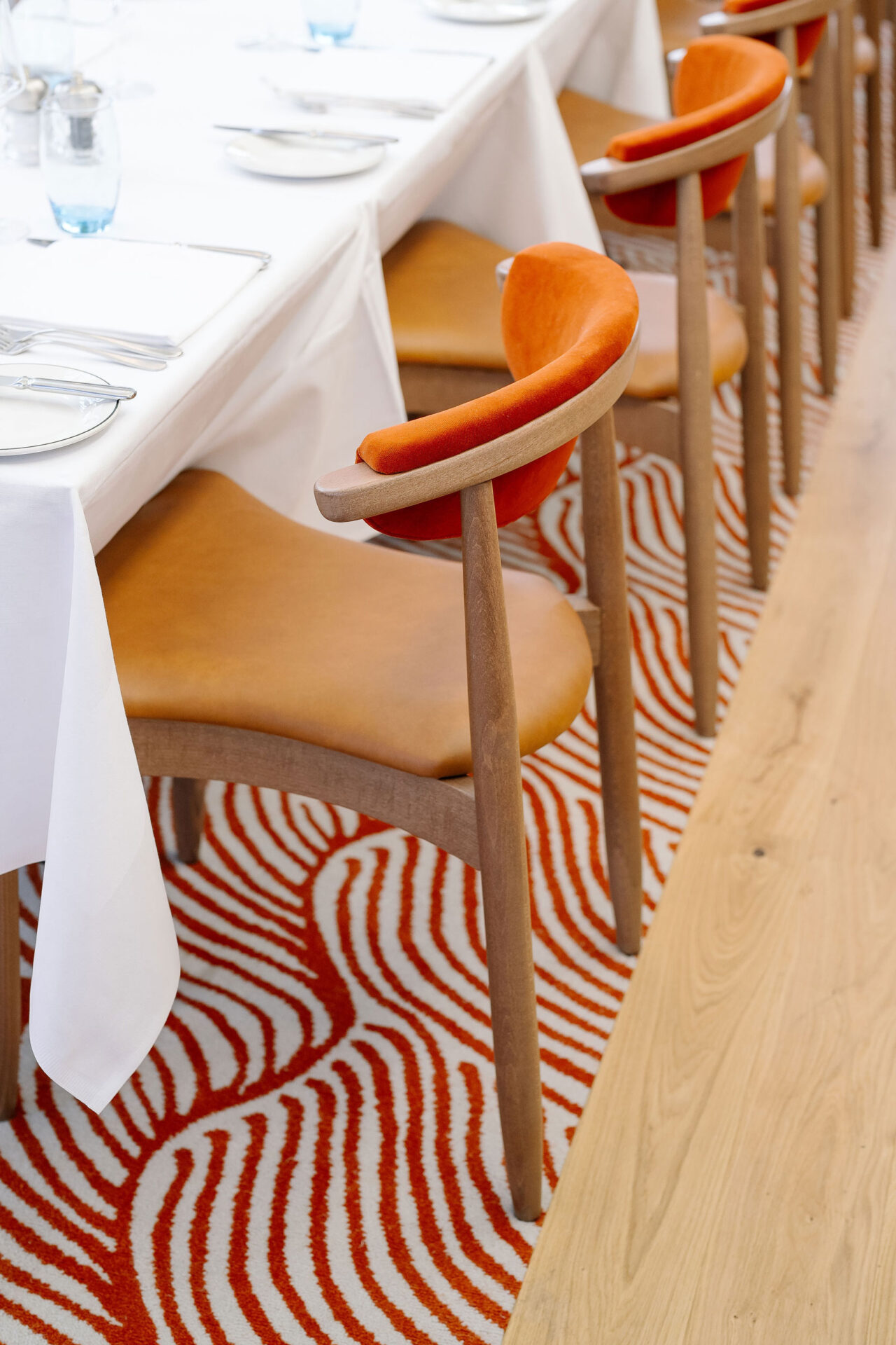 Tan leather and orange velvet upholstered wooden chairs facing a white tablecloth dining table, featuring a custom made lineart decorative rug inside Roast Restaurant, Borough Market London