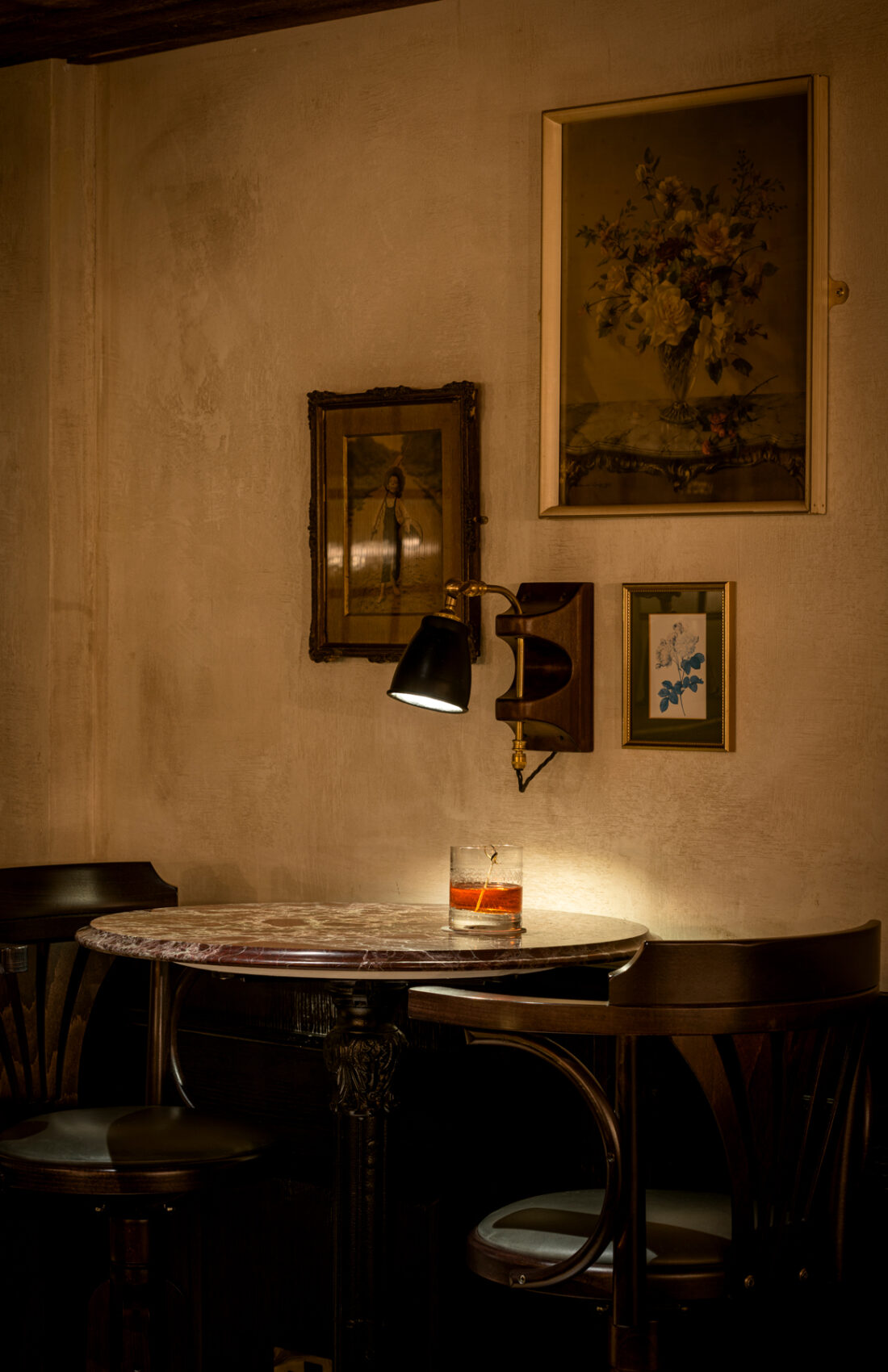 Moody, atmospheric, dim lit interior scene featuring vintage marble top table and old fashioned cocktail, next to wooden, leather upholstered chairs. On the walls there are vintage, reclaimed paintings. Inside Good's Way music venue and events space in King's Cross, London, designed by 3Stories Interior Design and Branding creative agency.