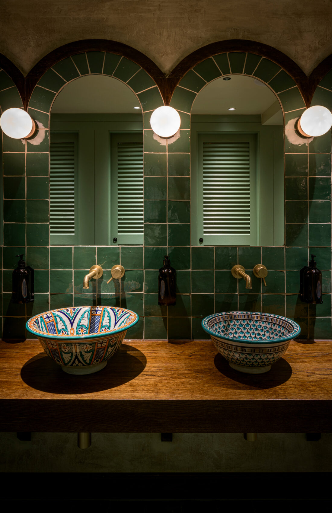 Green bathroom toilet tiles with arched mirrors, reclaimed hand painted porcelain sinks on wooden table top inside Good's Way music venue and events space in King's Cross, London, designed by 3Stories Interior Design and Branding creative agency.